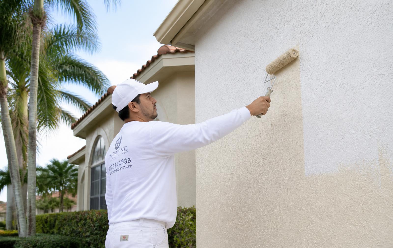 Painter in a white uniform rolling fresh off-white paint onto the stucco exterior wall of a South Florida home